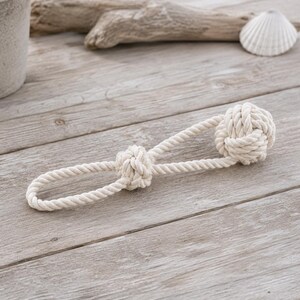 May include: A natural off-white cotton rope toy featuring a large monkey fist knot at one end and a smaller decorative knot securing a loop at the other end. The twisted rope construction creates a durable, textured surface for this pet accessory.
