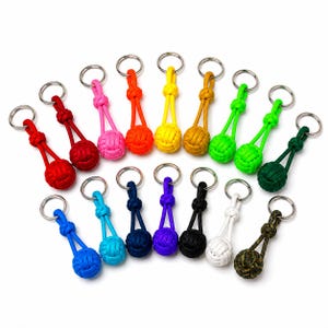 May include: A collection of sixteen monkey fist knot keychains made from paracord in various solid colors including red, pink, orange, yellow, gold, lime green, dark green, blue, cyan, navy, purple, black, white, and a camouflage pattern.