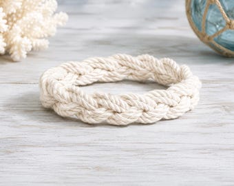 May include: A circular bracelet crafted from thick, off-white braided cotton rope. The design features a continuous, interwoven pattern created by multiple strands of rope, resulting in a textured, nautical-inspired accessory with a sturdy construction.