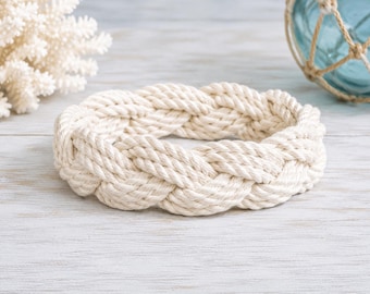 Op de afbeelding: Een circulair decoratief object gemaakt van dik, gebroken wit gevlochten touw. Het touw is geweven in een doorlopend, herhalend patroon dat een stevige, ronde ringvorm vormt met een getextureerd oppervlak en een natuurlijke uitstraling.