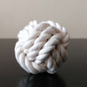 May include: A white cotton rope ball, knotted into a sphere shape. The ball is made of thick, braided rope and is designed for playing fetch or tug-of-war with dogs.