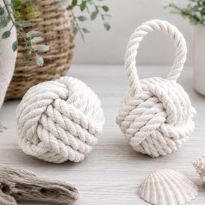 May include: Two off-white monkey fist knots made of thick twisted cotton rope. One knot is a spherical ball, while the other knot features an integrated rope loop handle extending from the top, both showcasing a complex, interwoven nautical design.