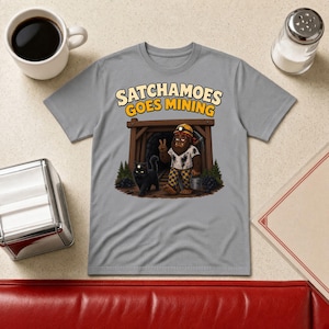May include: A grey t-shirt featuring a graphic of a brown creature wearing a miner's helmet and checkered pants, standing in a mine entrance with a black cat. The text above reads SATCHAMOES GOES MINING in bold, light-colored lettering.
