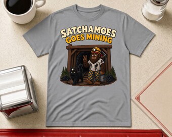 May include: A grey t-shirt featuring a graphic of a brown creature wearing a miner's helmet and checkered pants, standing in a mine entrance with a black cat. The text above reads SATCHAMOES GOES MINING in bold, light-colored lettering.