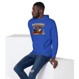 Puede incluir: Una sudadera con capucha de color azul real que presenta un estampado gr&aacute;fico de un personaje de dibujos animados en un kayak rojo con un gato negro. El texto sobre la ilustraci&oacute;n dice SATCHAMOES GOES KAYAKING en letras amarillas.
