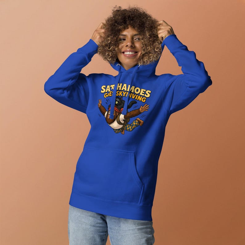 May include: A royal blue hooded sweatshirt featuring a graphic print of a brown creature skydiving with a black cat on its head. The text above the illustration reads "SATCHAMOES GOES SKYDIVING" in yellow block letters. The hoodie has long sleeves.