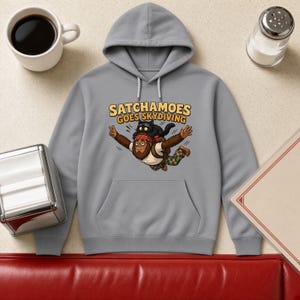 May include: A grey hooded sweatshirt featuring a graphic print of a brown creature with a black cat on its head, both skydiving. The text above the illustration reads SATCHAMOES GOES SKYDIVING in yellow letters with a dark outline.