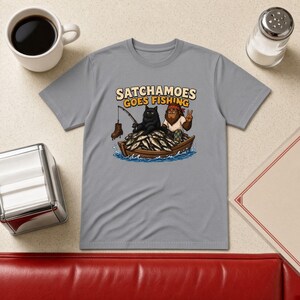 以下が含まれることがあります： グレーのTシャツで、魚でいっぱいの木製のボートに乗った黒猫と毛むくじゃらの生き物のグラフィックが描かれています。上部には「SATCHAMOES GOES FISHING」という文字があり、猫がブーツのついた釣り竿を持っている様子が描かれています。