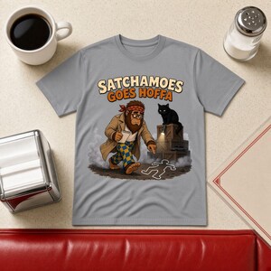 May include: A grey t-shirt featuring a cartoon graphic of a Bigfoot character wearing a tan trench coat and blue and yellow checkered pants, holding a notepad. Above, the text reads SATCHAMOES GOES HOFFA. A black cat with a flashlight stands on wooden crates nearby.