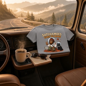 Puede incluir: Una camiseta gris con un gr&aacute;fico de un sasquatch de dibujos animados en traje espacial y un gato negro con casco espacial en Marte, con el texto SATCHAMOES GOES TO MARS, un gesto de paz y una peque&ntilde;a bandera con la palabra PEACE.