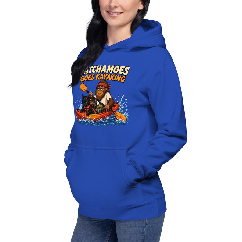 Puede incluir: Una sudadera con capucha azul real que presenta un estampado gr&aacute;fico de una criatura marr&oacute;n con un pa&ntilde;uelo rojo y una camisa blanca, remando en un kayak naranja con un gato negro. El texto sobre la ilustraci&oacute;n dice "SATCHAMOES GOES KAYAKING".
