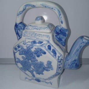 May include: A white ceramic teapot with a blue floral pattern and a geometric border design. The teapot features a curved handle, a short spout, and a removable lid with a small knob. The body is hexagonal with painted blue flowers.