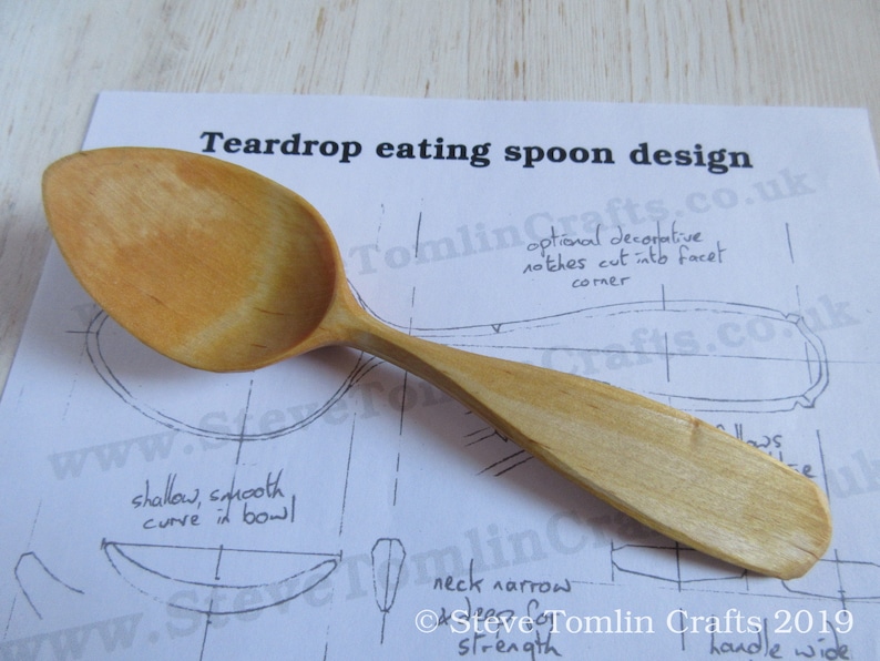 May include: A wooden teardrop-shaped spoon with a wide handle. The spoon is resting on a piece of paper with a drawing of the spoon and instructions for making it. The text on the paper reads "Teardrop eating spoon design" and "Steve Tomlin Crafts 2019".