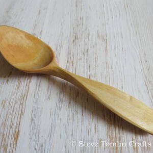 May include: A wooden spoon with a light brown color and a smooth, rounded surface. The spoon is made of natural wood and has a long handle.