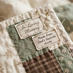 May include: A quilted fabric piece featuring two rectangular labels stitched onto the surface. The top label reads In Loving Memory, and the bottom label reads Made from Dad's favorite shirts. The quilt has cream, sage green, and brown squares.