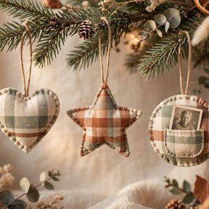 May include: Three fabric ornaments hanging from a pine branch. One is heart-shaped, one is star-shaped, and one is circular with a small pocket containing a black and white photograph of a person. All feature a green and brown plaid pattern.