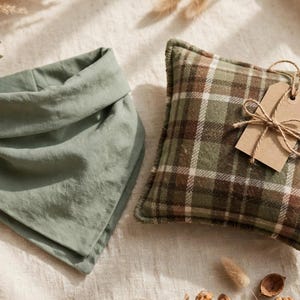 May include: A solid sage green triangular bandana next to a square pillow featuring a brown, green, and white plaid pattern. The pillow is adorned with a blank rectangular brown paper tag tied with a thin piece of natural twine.