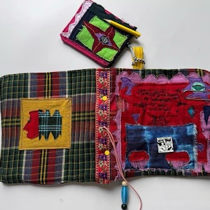 May include: A fabric journal cover featuring a green and red plaid exterior with a yellow patch and a red interior with blue and pink fabric appliqués, a floral trim, yellow buttons, a dangling bead, and a small matching zippered pouch.