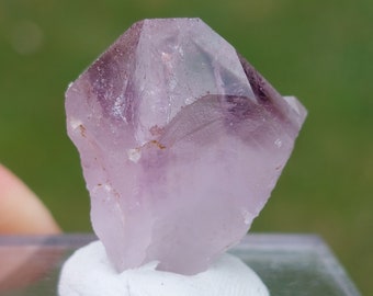 May include: A light purple amethyst crystal with a pointed tip and a clear, glassy appearance. The crystal is sitting on a white display stand.