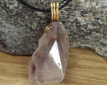 May include: A natural amethyst crystal pendant with a gold-colored wire wrap and a black leather cord. The crystal is a pale purple color with a pointed tip and a rough, textured surface. The pendant is displayed on a wooden surface.