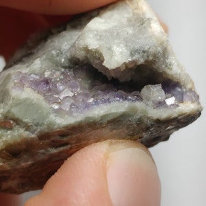 May include: A small, grey rock with a cavity filled with purple crystals. The crystals are clustered together and have a shiny, translucent appearance.
