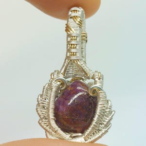 May include: A handcrafted pendant featuring a polished, oval-shaped, deep purple gemstone. The stone is encased in a woven wire setting of silver and gold-colored metal, with a loop at the top for a chain. The pendant is designed to be worn as jewelry.