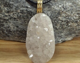 May include: A light gray, oval-shaped crystal pendant with a gold-colored bail, suspended from a black cord. The crystal has a rough, textured surface with many small, sparkling facets. The pendant is displayed against a gray stone background.
