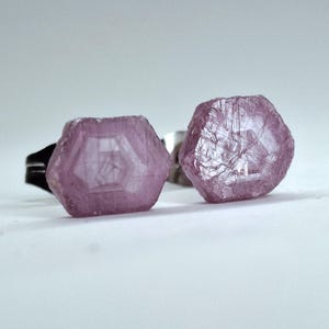 Puede incluir: Dos anillos con gemas rosas hexagonales en bruto. Las gemas tienen una superficie rugosa y texturizada, engastadas en bandas de metal plateado y negro. Los anillos se exhiben sobre una superficie blanca.