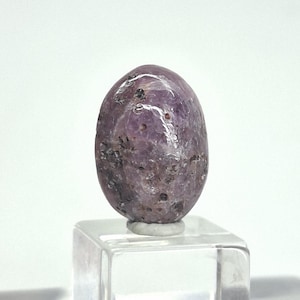 May include: A polished, oval-shaped gemstone in shades of purple and gray, resting on a clear, square display stand. The stone has a smooth, glossy surface with natural inclusions and variations in color.