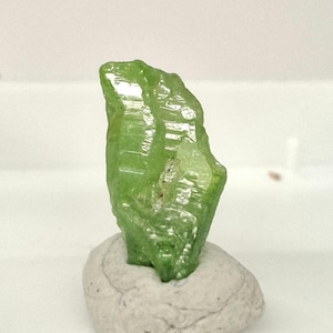 May include: A vibrant, translucent green crystal specimen. The gemstone is a light, bright green color with a rough, textured surface. It is mounted on a small, light gray, rounded stone base. The crystal is a natural mineral formation.