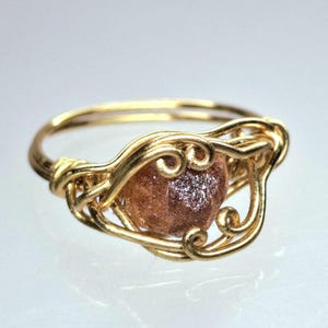 May include: A gold-colored wire-wrapped ring featuring a rough-cut, light brown gemstone. The ring's design includes intricate swirls and a double band, creating a unique and artistic piece of jewelry.