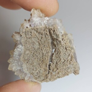 May include: A cluster of small, clear quartz crystals with a light purple hue, attached to a rough, gray, and brown rock. The crystals are clustered together in a jagged formation.