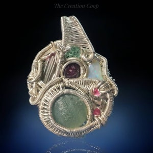 Puede incluir: Un colgante de alambre plateado envuelto alrededor de varias piedras preciosas, incluyendo verde, rosa y blanco. El colgante es una pieza de joyería única e intrincada.