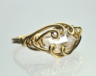 May include: A gold-colored wire-wrapped ring featuring a clear, faceted crystal. The ring's design incorporates intricate swirls and curves around the crystal, creating a delicate and artistic appearance. The ring is set against a reflective surface.