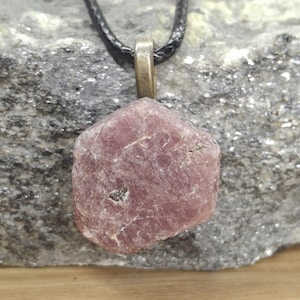 May include: A raw, pink gemstone pendant with a textured surface. The pendant is suspended from a black cord with a gold-colored metal clasp. The gemstone is set against a gray, textured background.