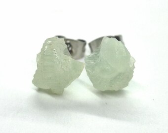 May include: A pair of light green, raw gemstone stud earrings. The earrings have a rough, textured surface and are set on silver-colored posts. The background is plain white.