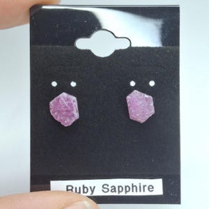 Puede incluir: Un par de pendientes de rubí zafiro. Cada pendiente presenta una gema rosa hexagonal de corte rugoso. Los pendientes se exhiben en una tarjeta negra con el texto "Ruby Sapphire" en blanco.