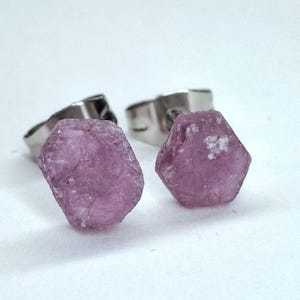 Puede incluir: Un par de pendientes de botón de piedras preciosas rosas en bruto. Las piedras preciosas tienen una textura natural rugosa y están engastadas en postes de color plateado. Los pendientes se muestran sobre un fondo blanco.
