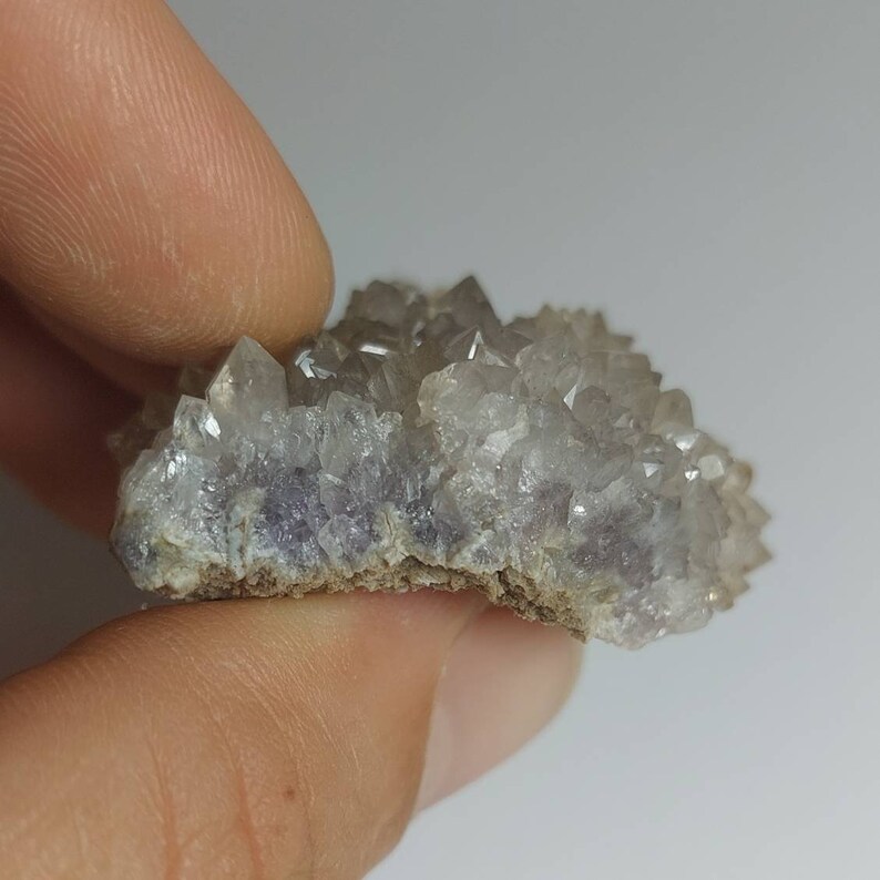 May include: A cluster of small, clear quartz crystals with a hint of purple, set on a rough, gray base.