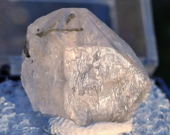 May include: A translucent, light-colored crystal with a rough, textured surface. The crystal has a slightly elongated shape and appears to be resting on a white base. Small, dark inclusions are visible within the crystal structure. The overall appearance suggests a natural mineral specimen.