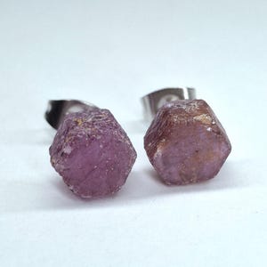 Op de afbeelding: Een paar ruwe robijn oorbellen. Elke oorbel heeft een gefacetteerde, ruw geslepen robijn edelsteen in roze en bruine tinten, gezet op een zilverkleurige metalen achterkant. De edelstenen hebben een natuurlijk, getextureerd uiterlijk tegen een witte achtergrond.