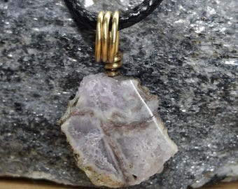 May include: A pendant necklace featuring a raw, irregularly shaped, light purple stone with brown veining. The stone is suspended from a gold-colored metal bail and a black braided cord. The necklace is displayed on a gray stone surface.
