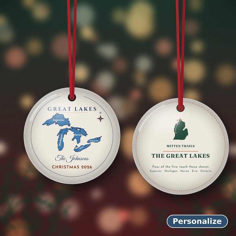 May include: Two round white ceramic ornaments with red ribbon hangers. The left ornament features a blue map of the Great Lakes, the text Great Lakes, The Johnsons, and Christmas 2026. The right ornament shows a green Michigan map, Mitten Trails, and text.