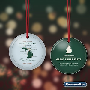 May include: Two round ornaments with red hanging ribbons. The left ornament is light blue with a map of Michigan, text reading New Home In Michigan, The Johnsons, Est. 2026. The right ornament is dark green with a white Michigan map, Mitten Trails, Great Lakes State, and text.