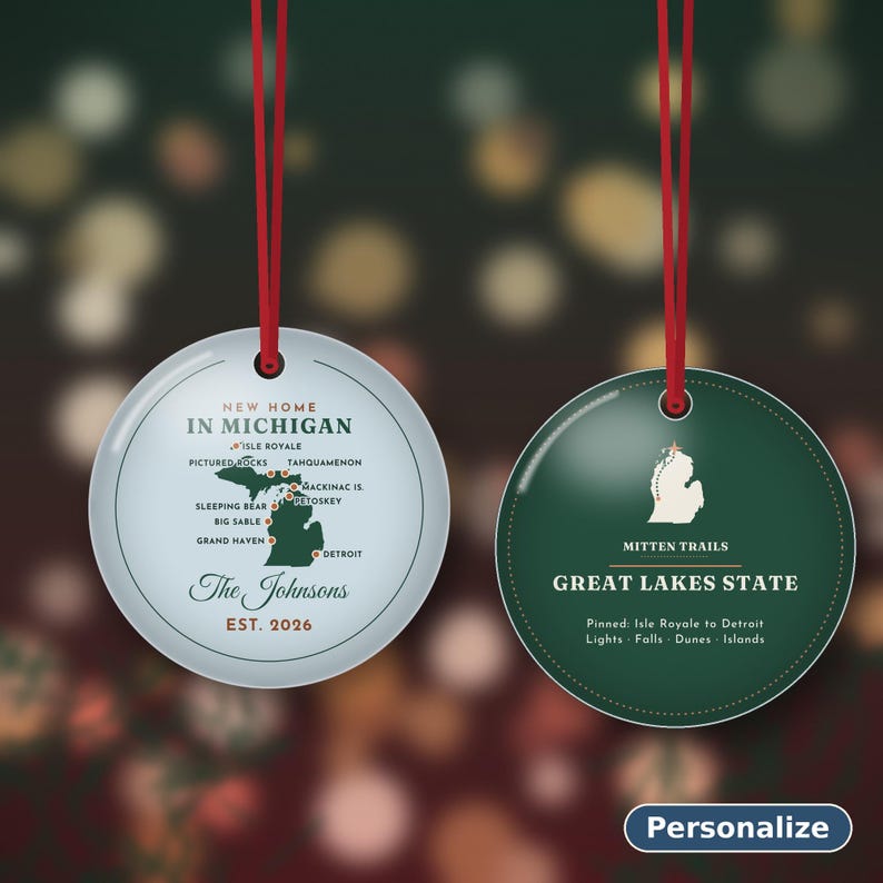 May include: Two round ornaments with red hanging ribbons. The left ornament is light blue with a map of Michigan, text reading New Home In Michigan, The Johnsons, Est. 2026. The right ornament is dark green with a white Michigan map, Mitten Trails, Great Lakes State, and text.