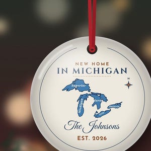May include: A round white ceramic ornament hanging from a red ribbon. The design features a blue map of the Great Lakes with the text NEW HOME IN MICHIGAN, The Johnsons, and EST. 2026. A small compass rose is positioned to the right of the map.