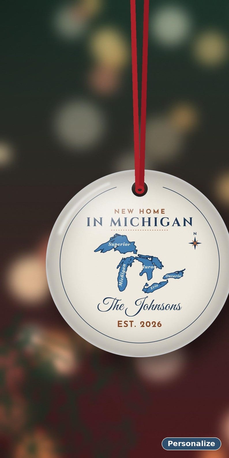 May include: A round white ceramic ornament hanging from a red ribbon. The design features a blue map of the Great Lakes with the text NEW HOME IN MICHIGAN, The Johnsons, and EST. 2026. A small compass rose is positioned to the right of the map.