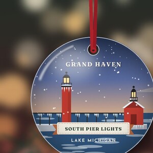 May include: A round, flat ornament hanging from a red ribbon, featuring a graphic illustration of the Grand Haven South Pier Lights on Lake Michigan. The design shows red lighthouses against a dark blue, snowy sky with white text.