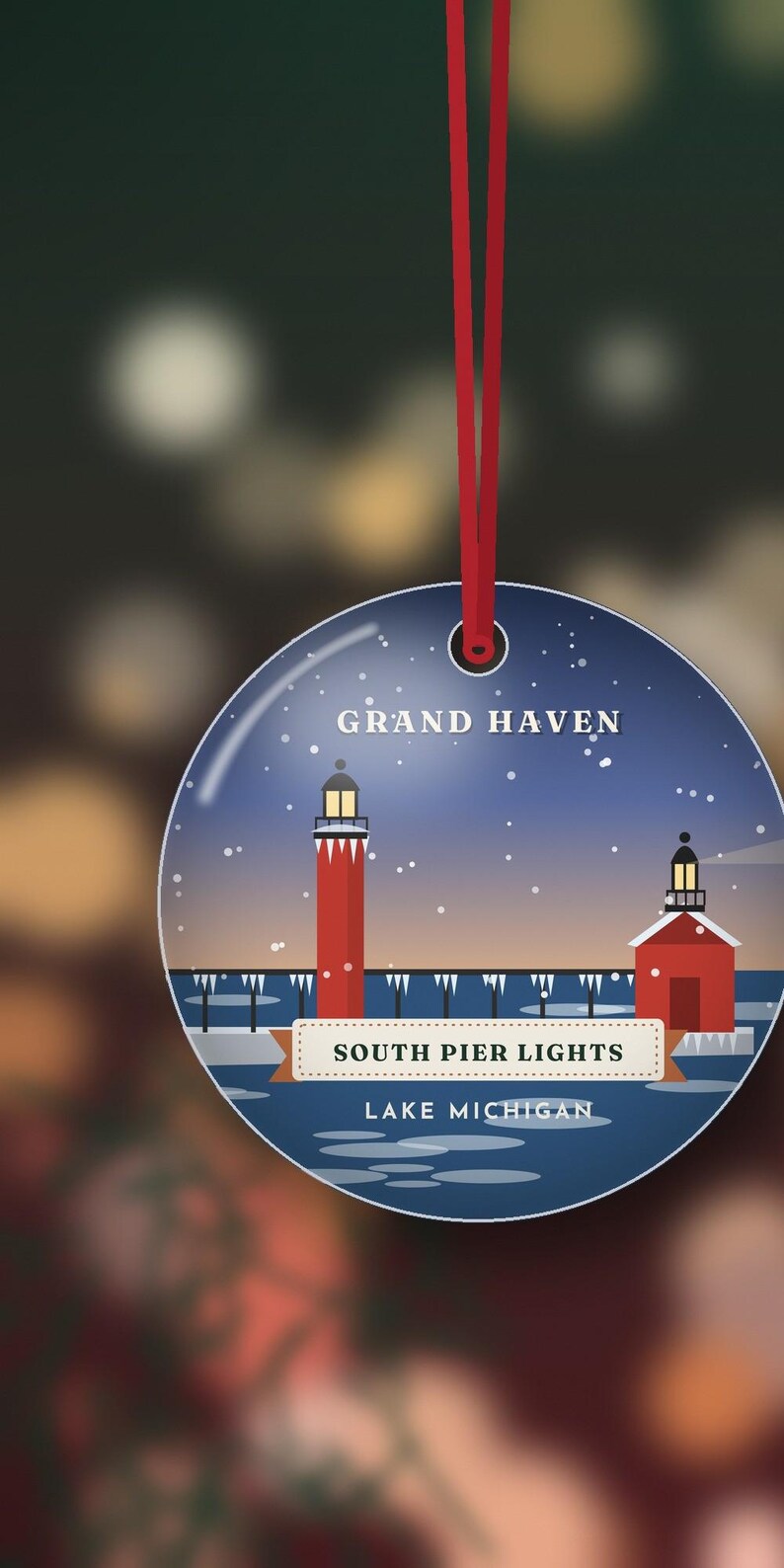 May include: A round, flat ornament hanging from a red ribbon, featuring a graphic illustration of the Grand Haven South Pier Lights on Lake Michigan. The design shows red lighthouses against a dark blue, snowy sky with white text.