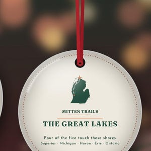 May include: A round white ornament hanging from a red ribbon. It features a green silhouette of Michigan with a star, the text MITTEN TRAILS, THE GREAT LAKES, and the words Four of the five touch these shores Superior Michigan Huron Erie Ontario.